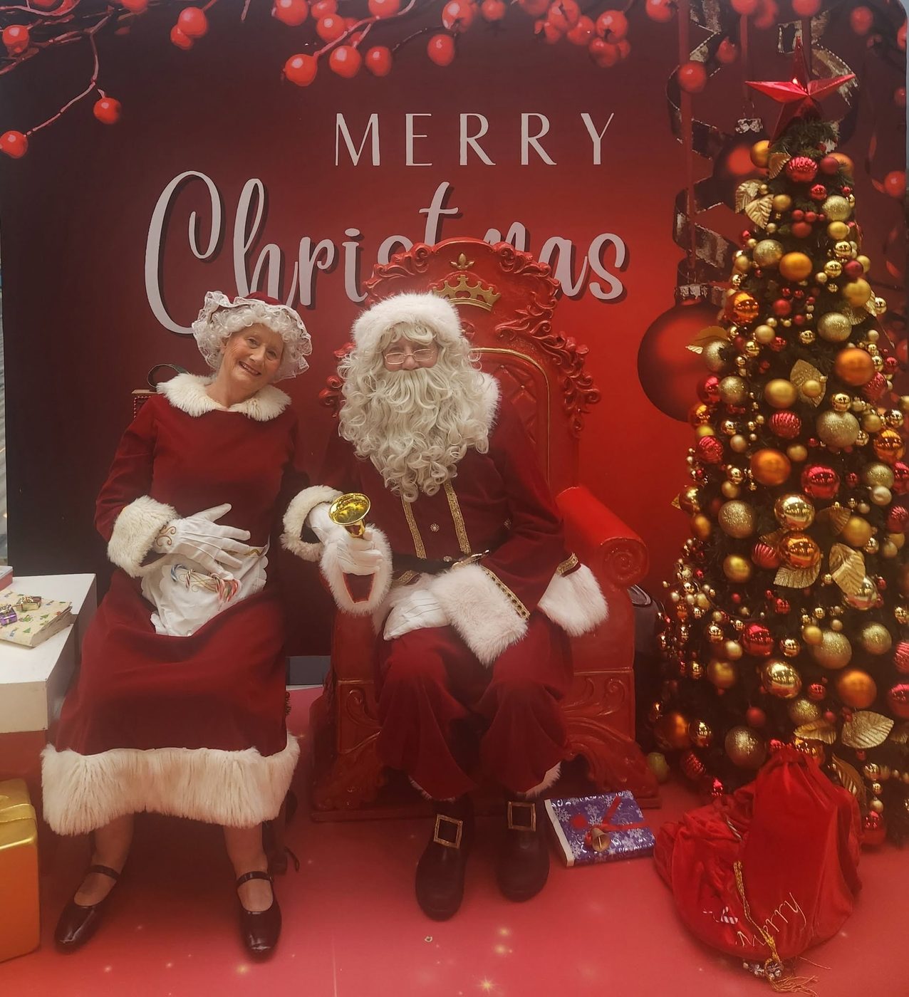Santa and Mrs Claus by a Merry Christmas backdrop